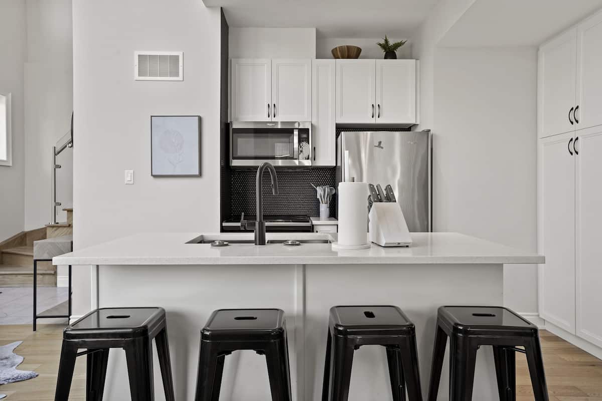 Una cocina moderna con armarios blancos, un refrigerador de acero inoxidable, un microondas y un lavamanos. Hay tres taburetes negros frente a la encimera.
