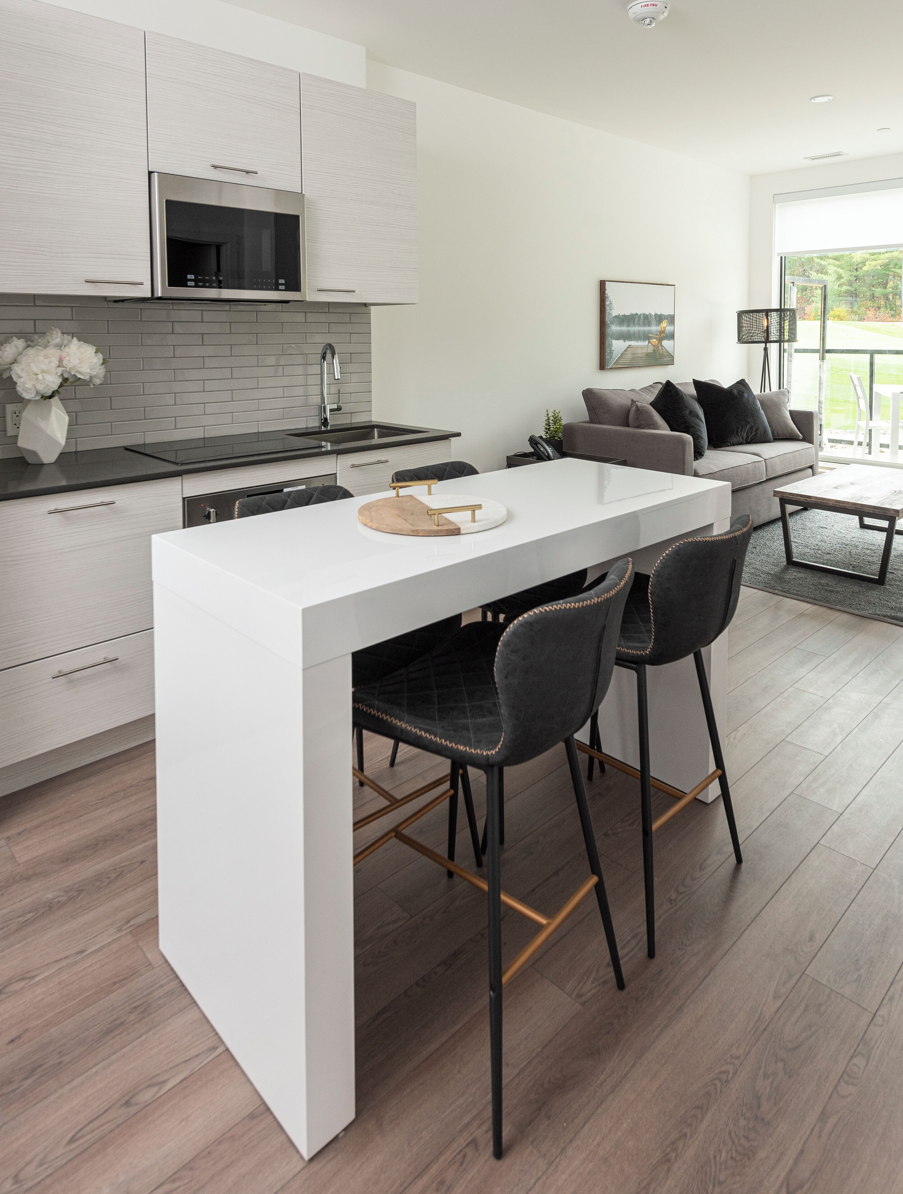 A modern kitchen with a white island, bar stools, and a grey sofa in a bright living area with a view.