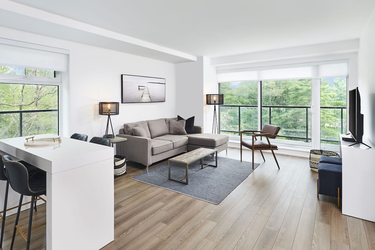 Una sala moderna con un sofá, una mesa de centro y sillas. Hay una zona de comedor con una mesa y sillas. Amplias ventanas ofrecen vistas al exterior.