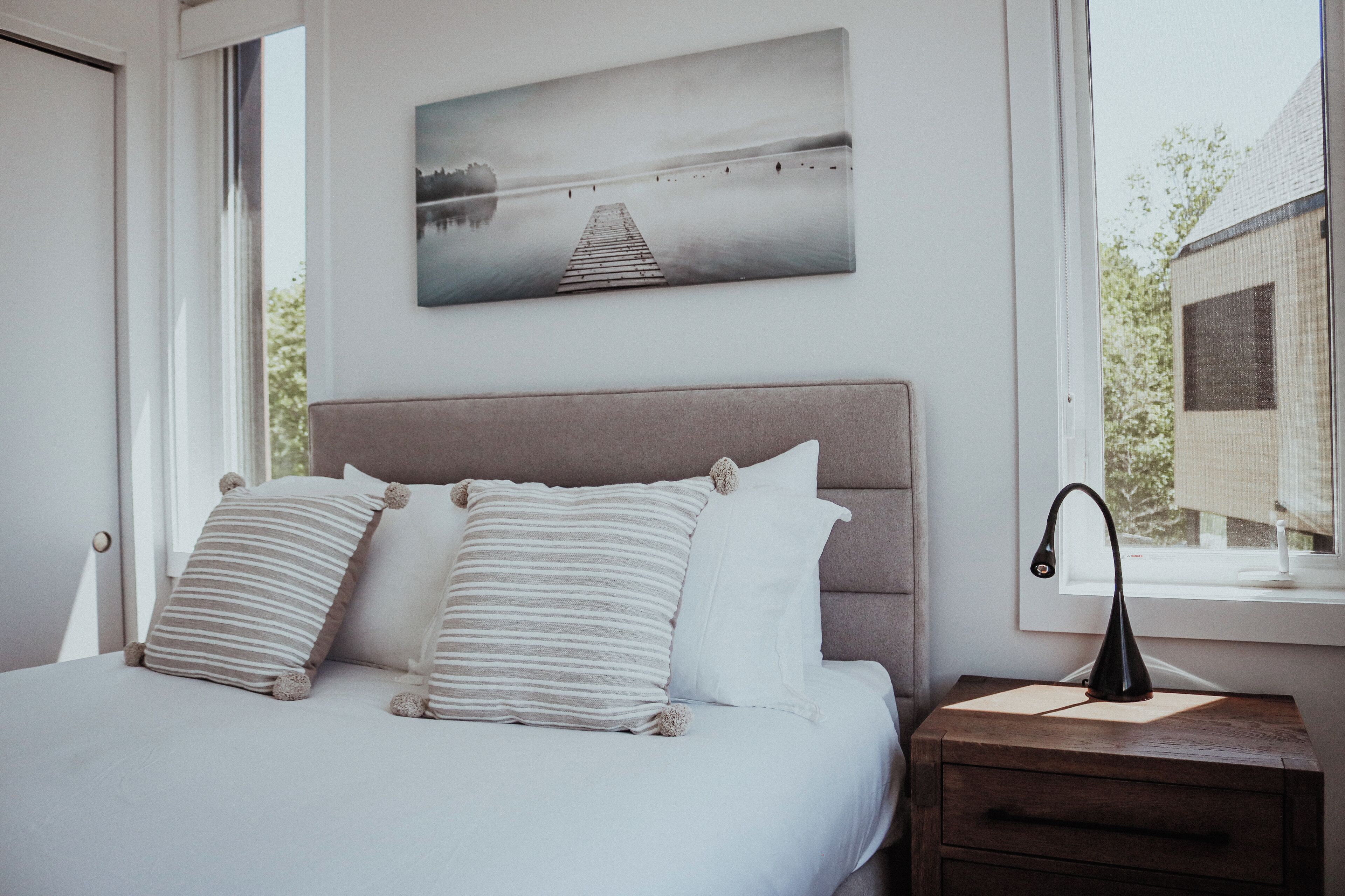 A bedroom with a bed, pillows, a nightstand, a lamp, a window, and a painting on the wall.