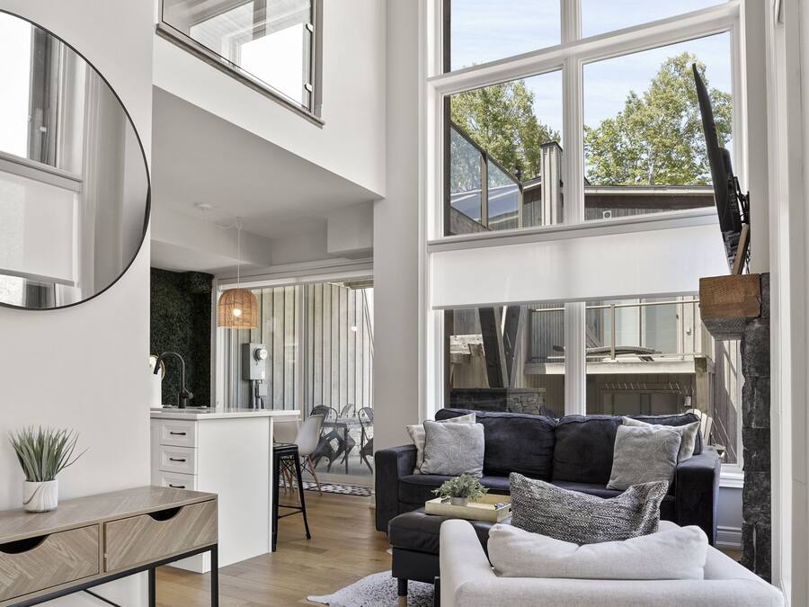 A spacious living area with a high ceiling, large windows, a modern kitchen, and a cozy seating area with a sofa and a coffee table.
