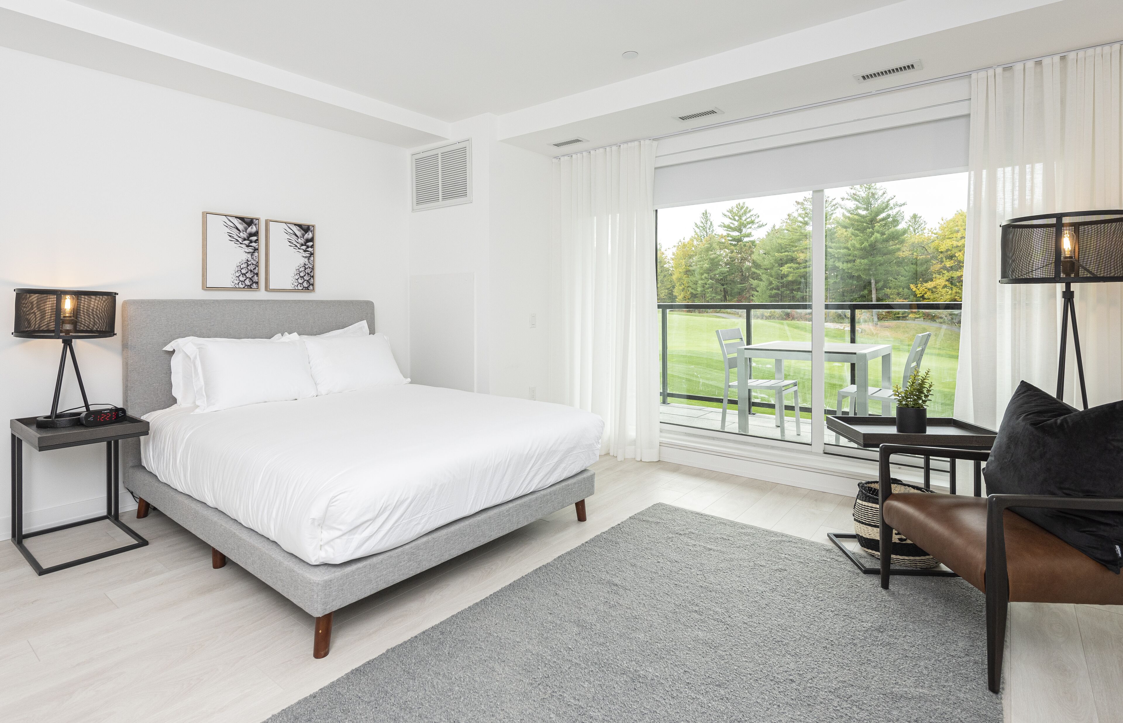 A modern bedroom with a large bed, a bedside table with a lamp, a view of a golf course through a sliding glass door, and a seating area with a table and chairs.