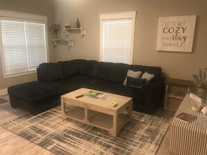 Una sala de estar con un sofá seccional oscuro, una mesa de centro de madera y una alfombra estampada. Hay dos ventanas con persianas, un estante montado en la pared con libros y una estatua, y un letrero que dice "Let's get cozy and stay awhile.".