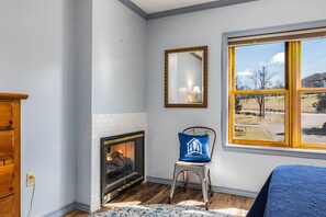A room with a fireplace, a chair with a cushion, a mirror, and a window with a view of the outdoors.