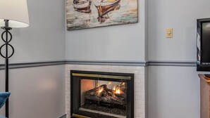 A fireplace with a white brick surround and a modern design, a painting of boats on the wall, and a television on the right. - Relaxing Suite Steps to Table Rock, Near Big Cedar, Community Hot Tub & Free Daily Tickets (Hollister)
