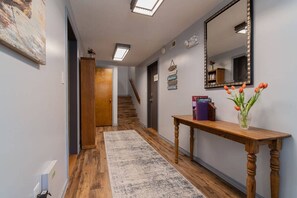 A hallway with a wooden floor, a rug, a wooden console table with a vase of flowers, a mirror, and a staircase in the background.