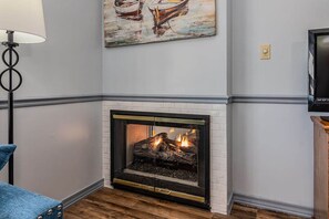 A fireplace with a white brick surround, a television mounted on the wall to the right, and a painting of boats above the fireplace.
