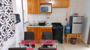 A compact kitchen with wooden cabinets, a microwave, a toaster, a blender, a stove, and a refrigerator. There is a dining table with chairs and a patterned curtain on the left.