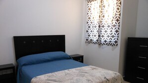 A bedroom with a bed, a nightstand, a dresser, and a window with patterned curtains.