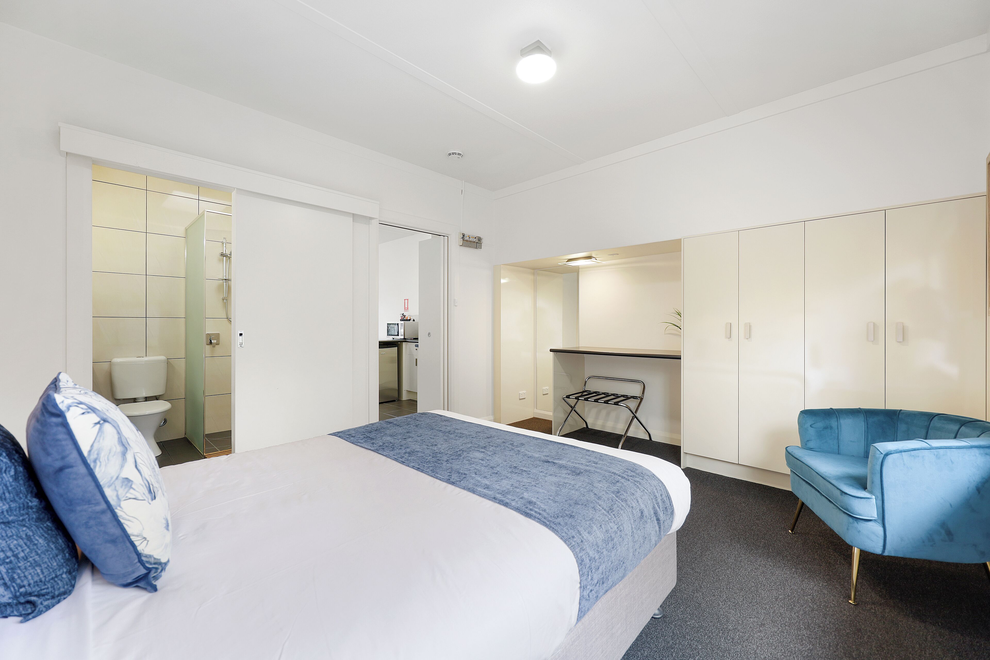 Ein Hotelzimmer mit Bett, blauem Sessel, Kleiderschrank und einem durchgegangenen Tür erkennbaren Badezimmer.