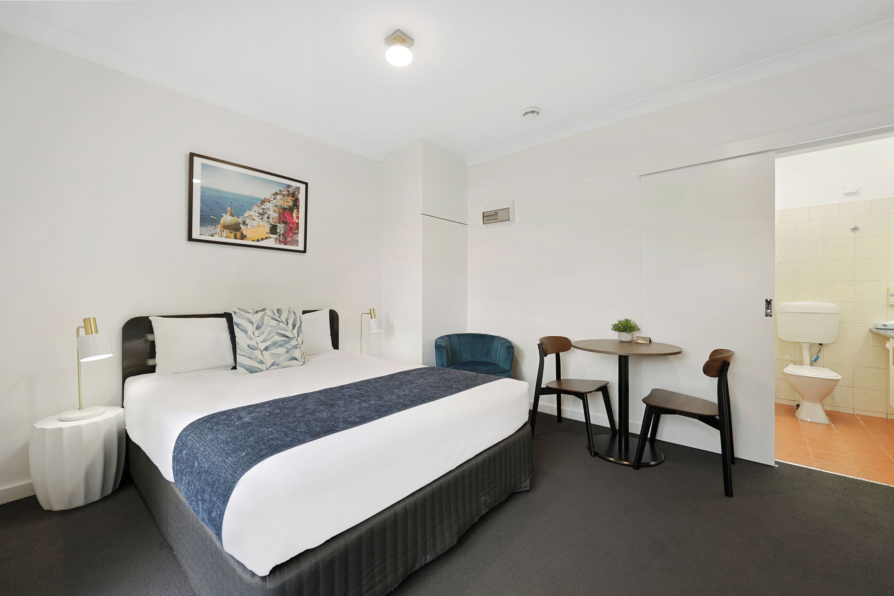 A hotel room with a large bed, a desk, two chairs, a small table with a plant, and a picture on the wall.