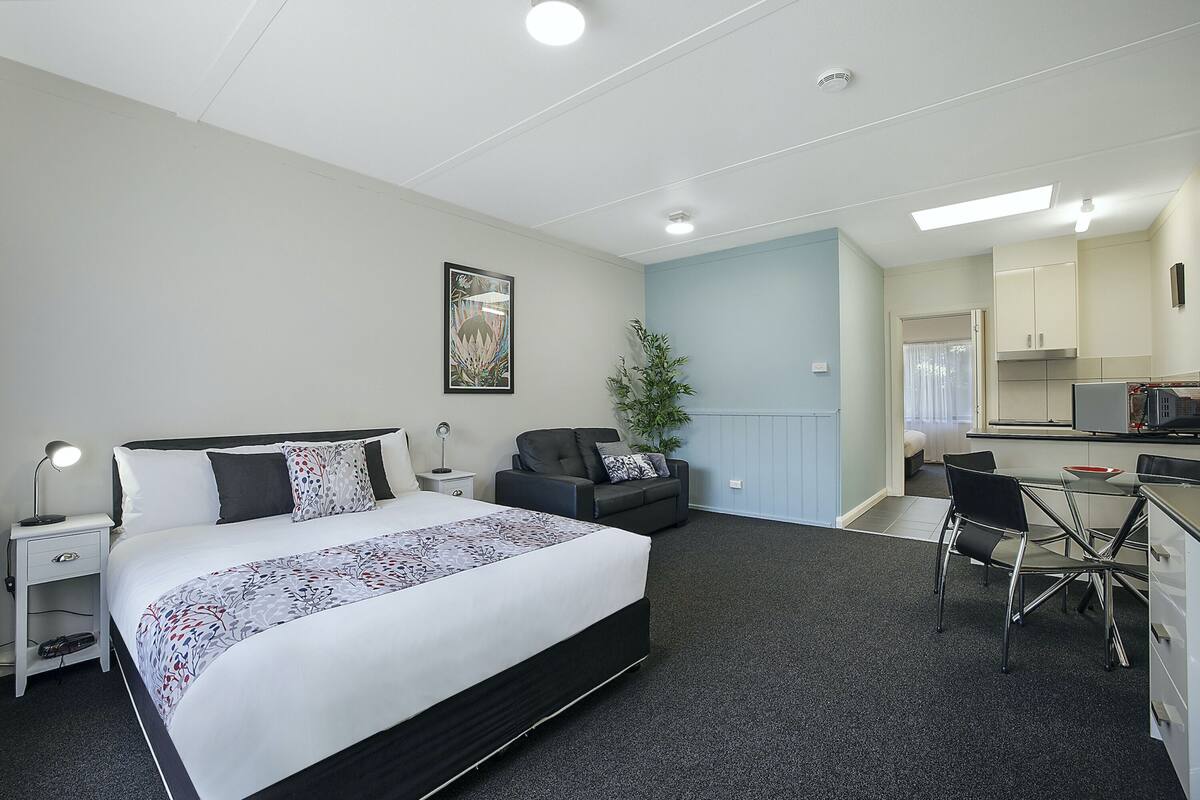 Quarto de hotel com cama, mesa de cabeceira, luminária, área de estar e cozinha compacta.