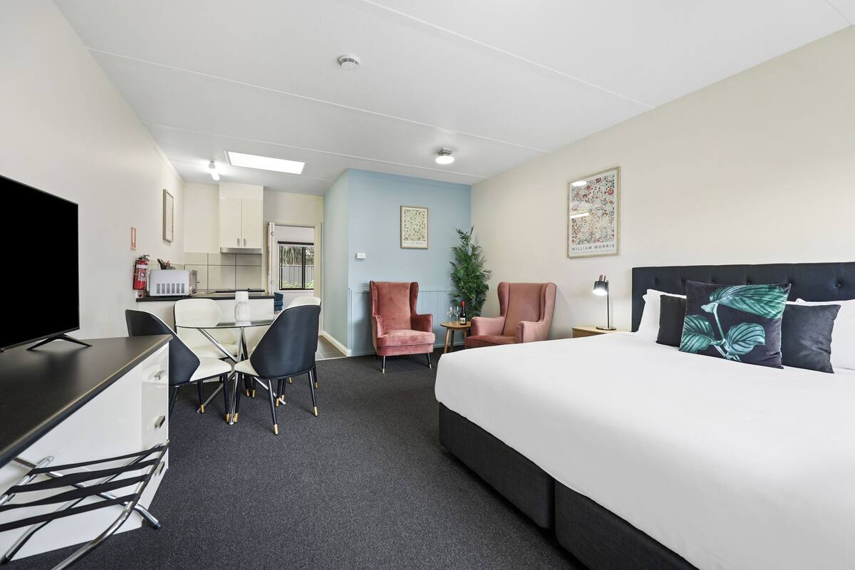 Quarto de hotel com cama, escrivaninha, cadeira, mesa pequena e planta em vaso.