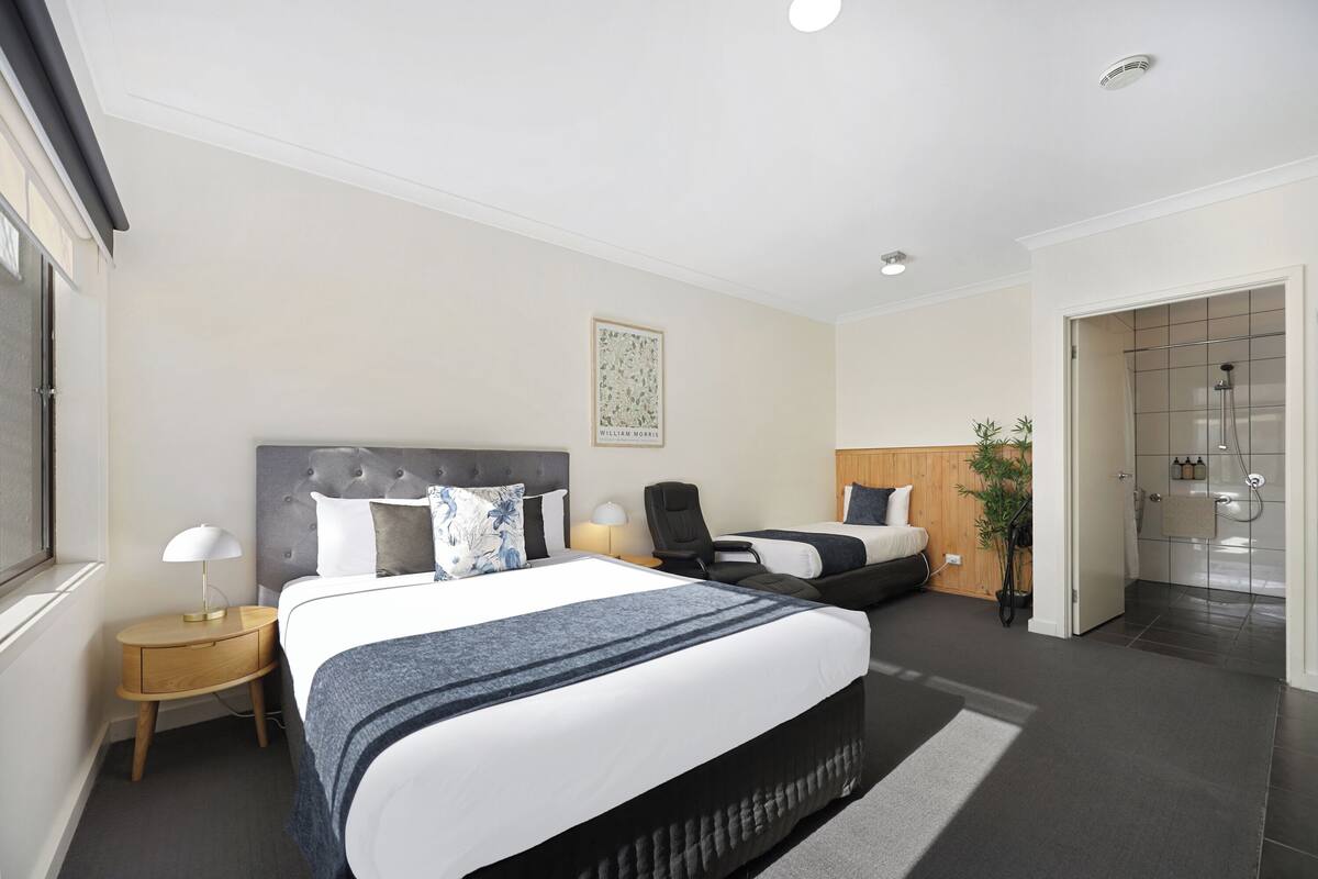 Quarto de hotel moderno com uma cama grande, uma mesinha de cabeceira, uma cadeira, uma escrivaninha, área de chuveiro e um quadro na parede.