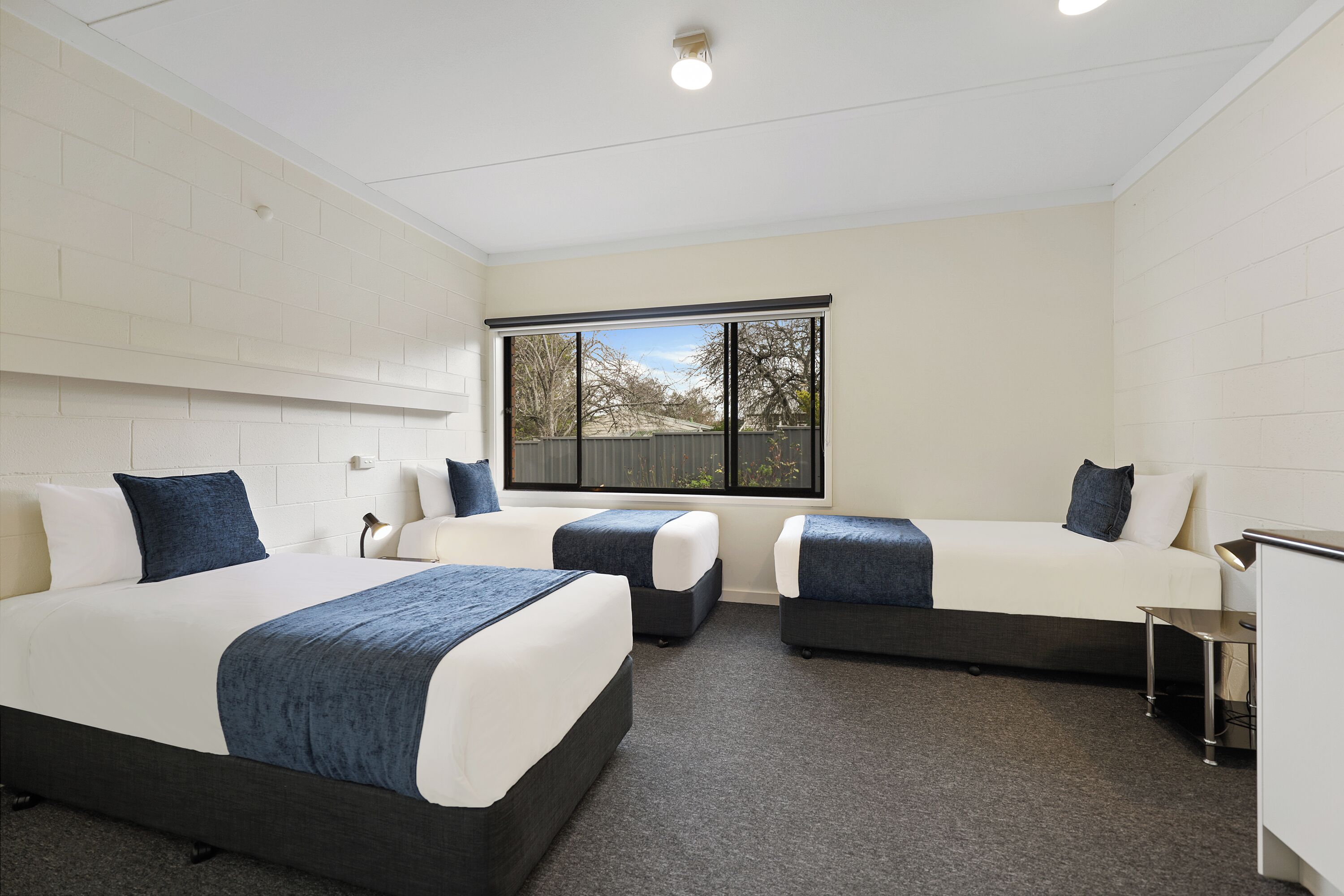 Ein Hotelzimmer mit drei Betten, einem Schreibtisch und einem großen Fenster mit Blick auf Bäume und einen Zaun.