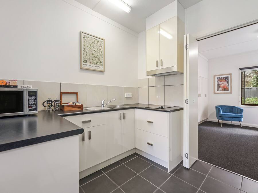 A modern kitchen with white cabinets, a microwave, and a sink. There is a framed artwork on the wall and a blue chair in the corner.