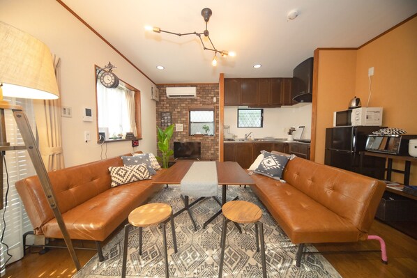 A cozy living room with a sofa, coffee table, and chairs, a kitchen area with cabinets and appliances, and a decorative ceiling light. - Kanegafuchi House (Sumida-ku)
