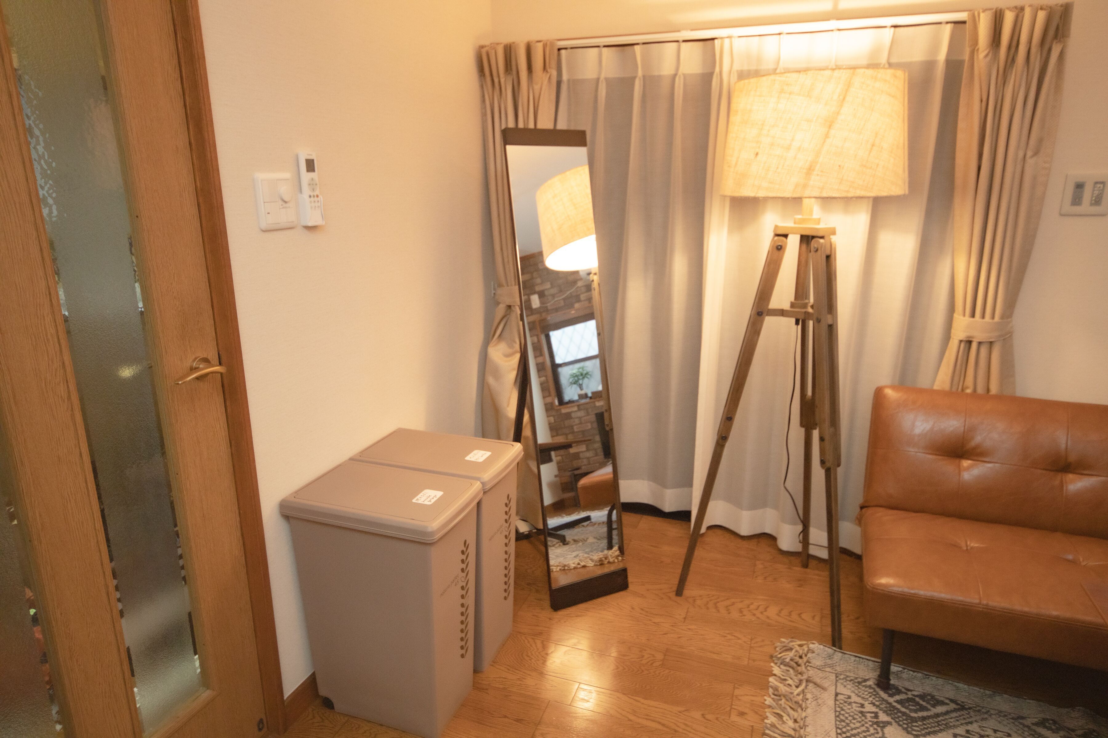 A room with a wooden floor, a brown leather sofa, a standing lamp, a mirror, and a small white cabinet.