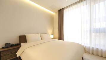 A hotel room with a large bed, a bedside table with a phone, and a window with sheer curtains.