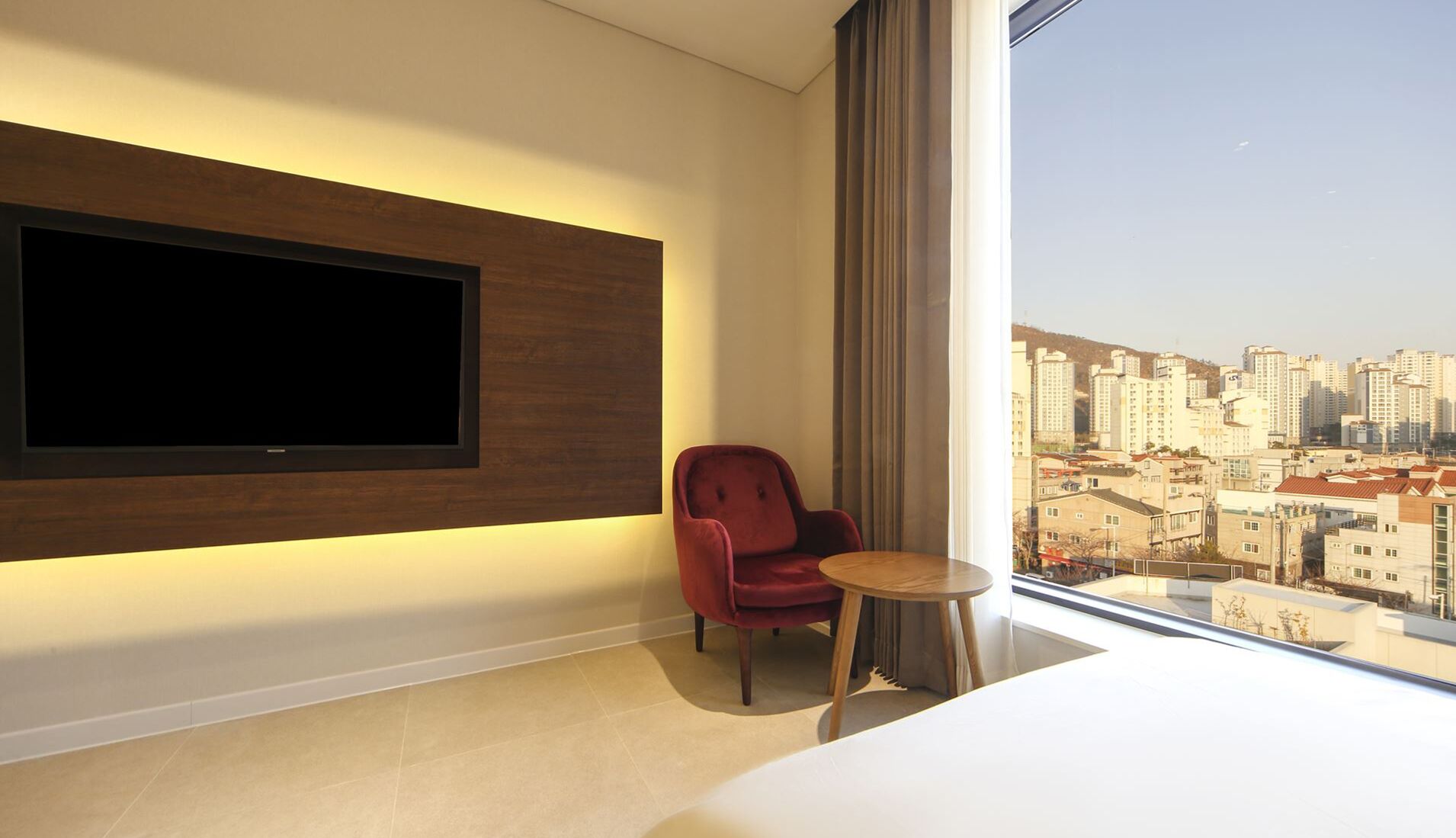 A modern hotel room with a large TV mounted on the wall, a red chair, a small round table, and a large window offering a city view.