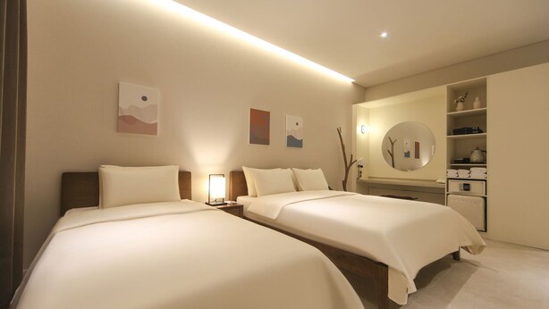 A hotel room with two beds, a bedside table with a lamp, a large mirror, and a wardrobe with shelves. - Hermon Hotel Gangseo (Busan)