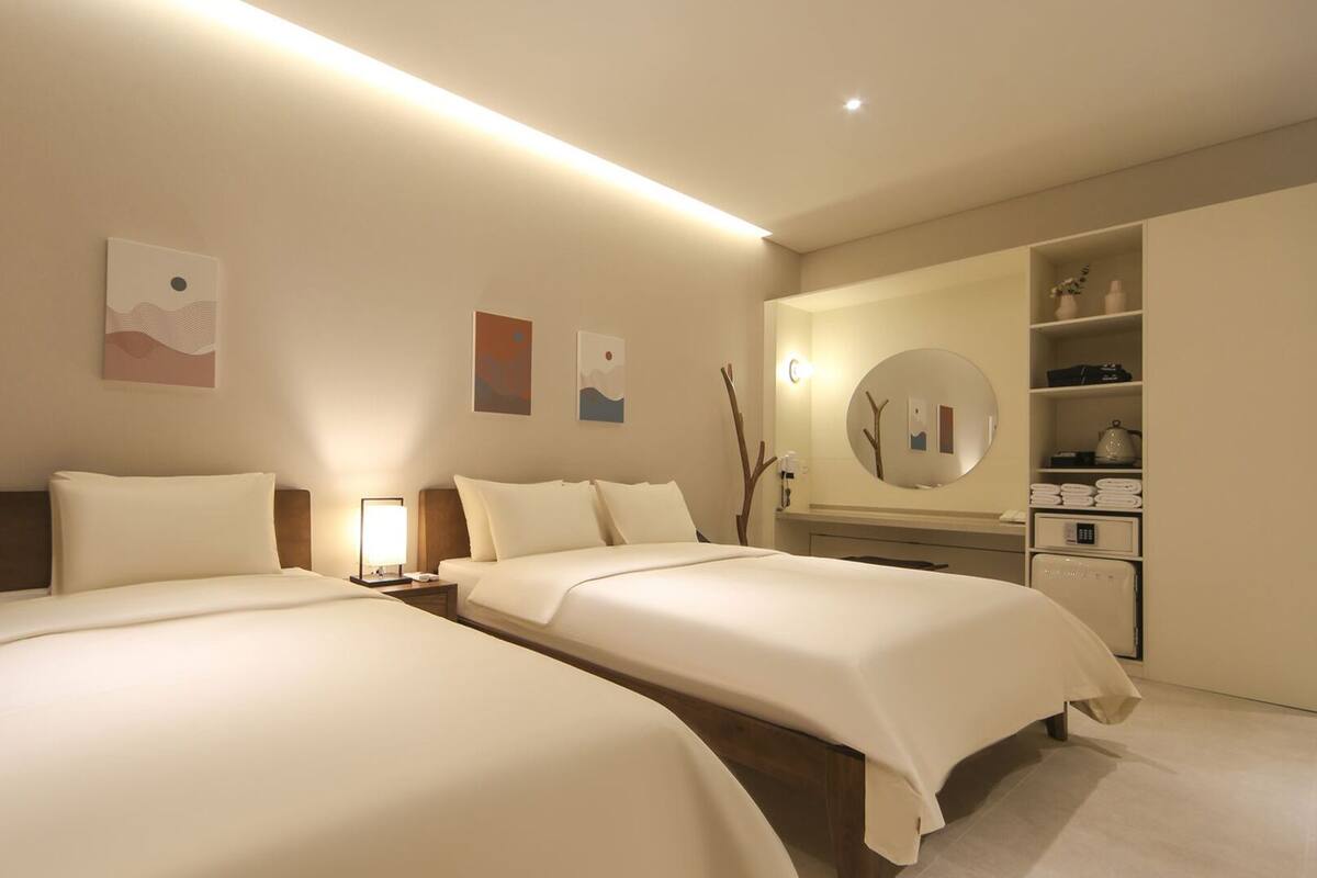A hotel room with two beds, a bedside table with a lamp, a large mirror, and a wardrobe with shelves.