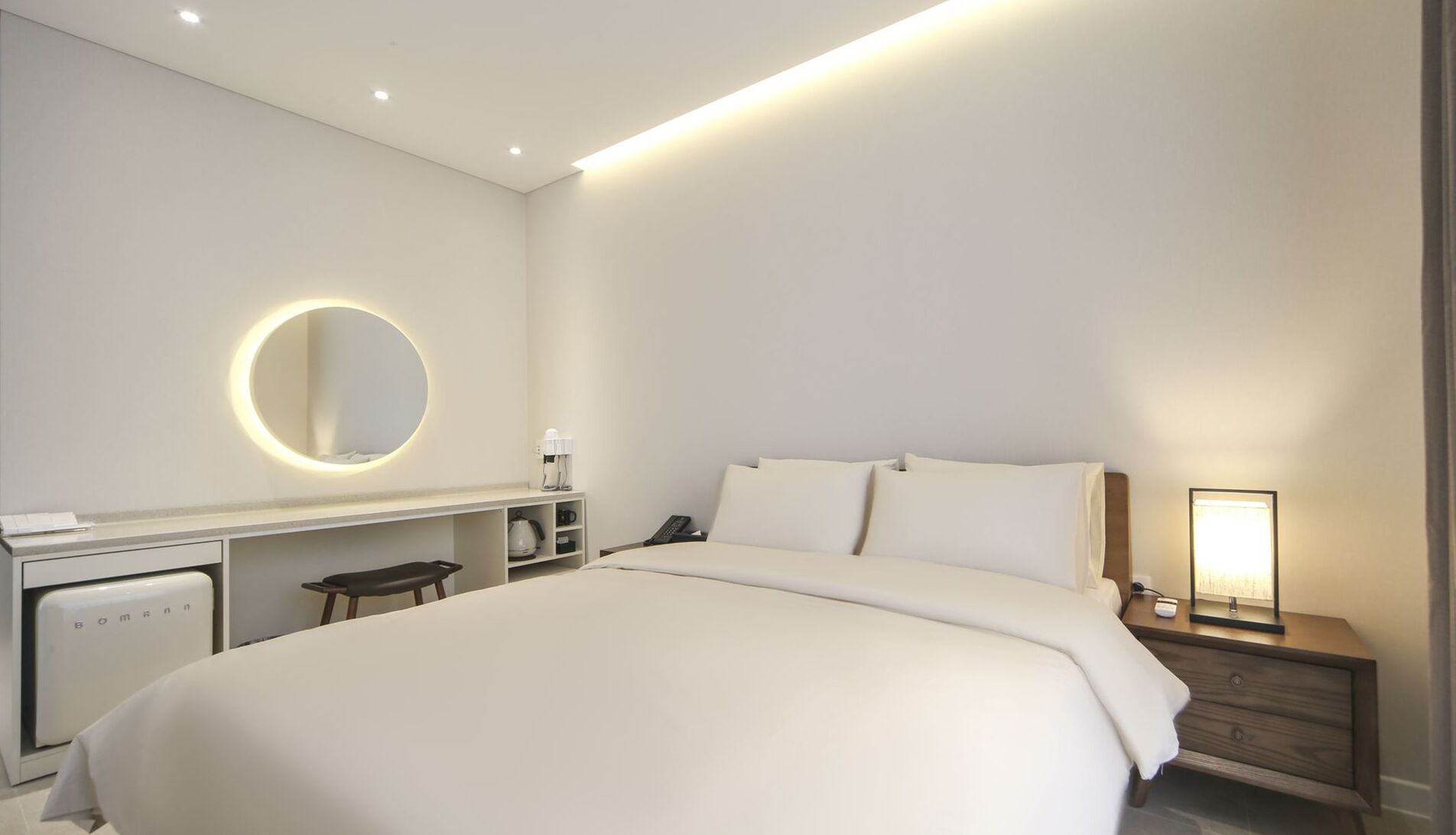 A modern hotel room with a large bed, a desk with a round mirror, a coffee machine, and a bedside table with a lamp.