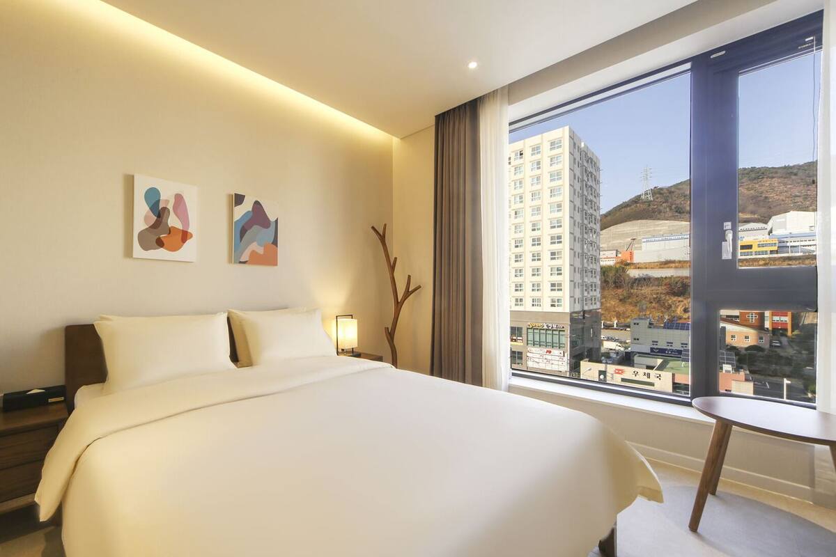 A hotel room with a large bed, a bedside table with a phone, a small table, and a window with a view of buildings and hills.