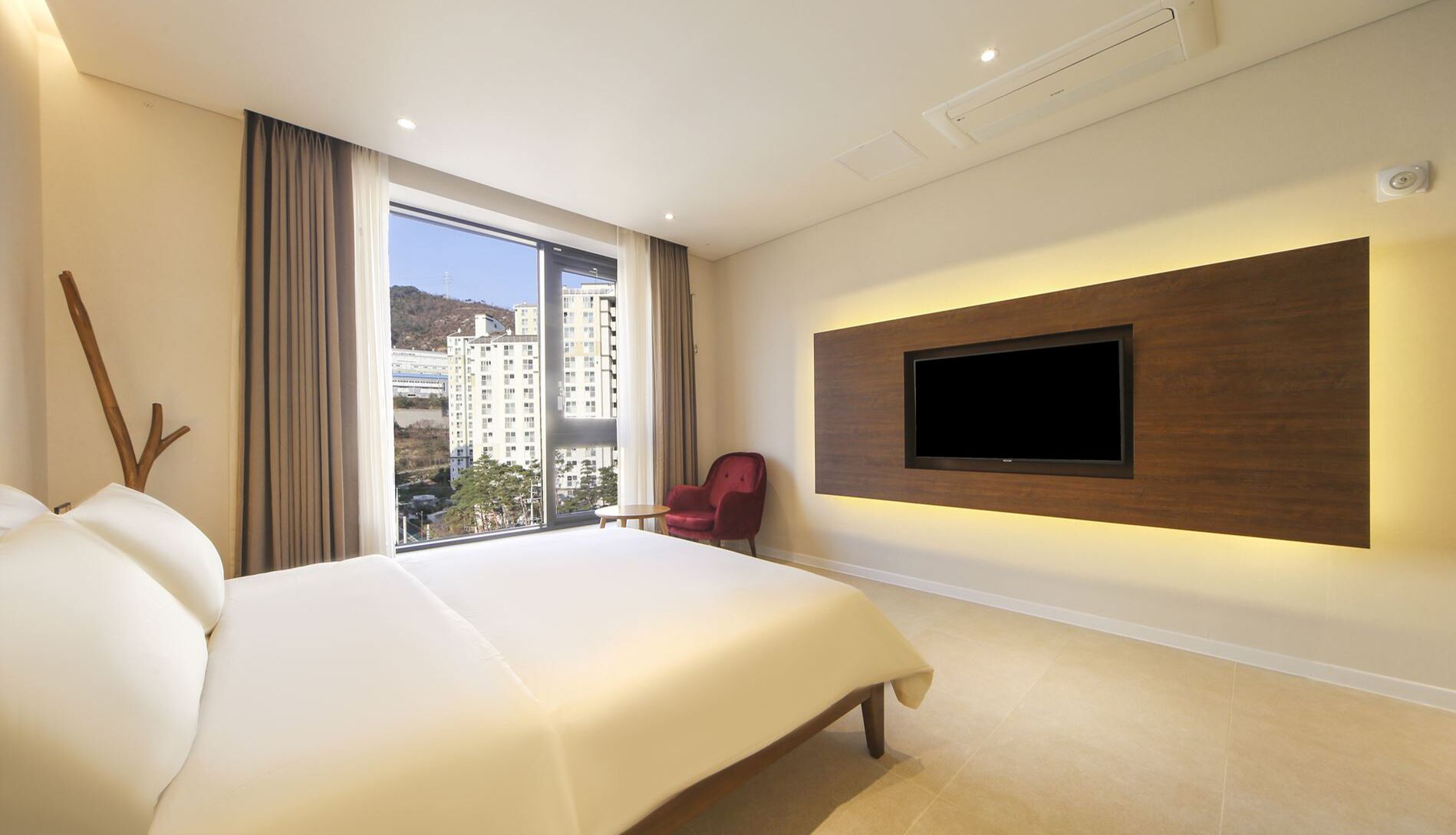 A hotel room with a large bed, a mounted TV, a red chair, and a view of buildings through the window.