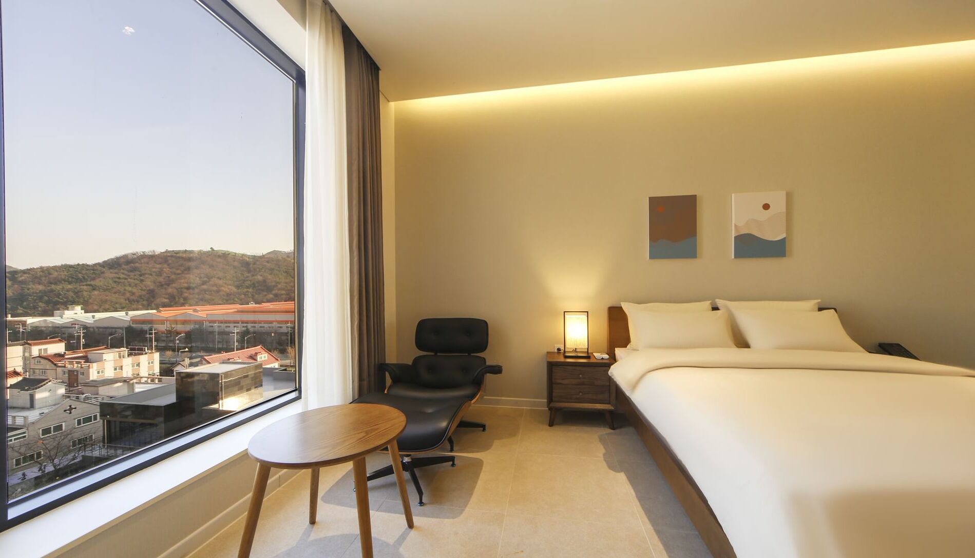 A hotel room with a large window, a bed, a chair, a nightstand, and two wall art pieces.