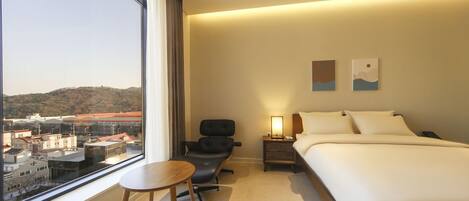 A hotel room with a large window, a bed, a chair, a nightstand, and two wall art pieces.