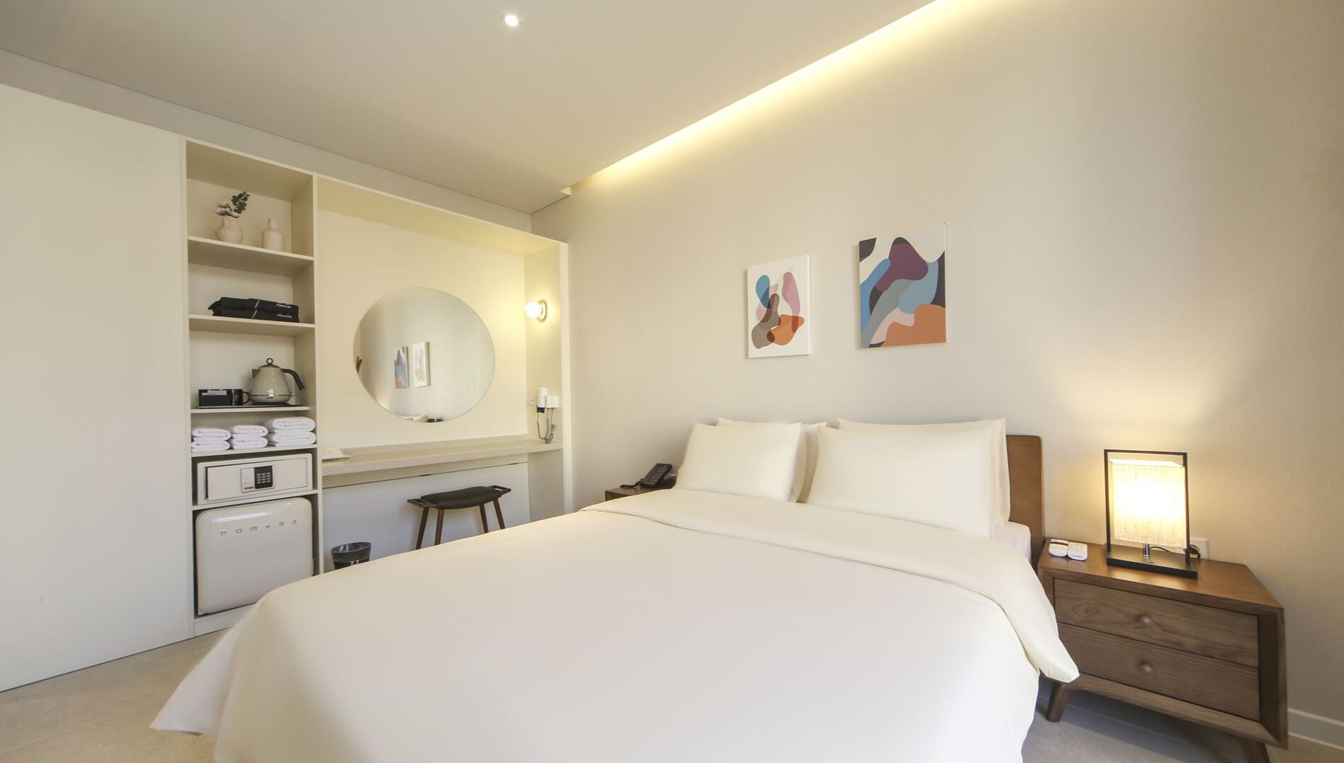 Une chambre d’hôtel avec un grand lit, un bureau avec une chaise, un miroir rond et deux tableaux abstraits accrochés au mur.