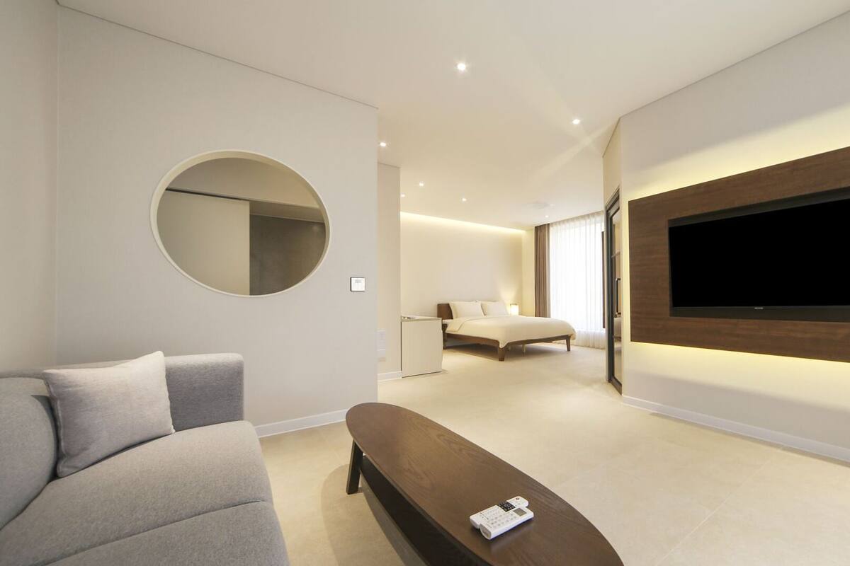 A modern hotel room with a large flat-screen TV mounted on the wall, a round mirror, a grey sofa, and a wooden bench.