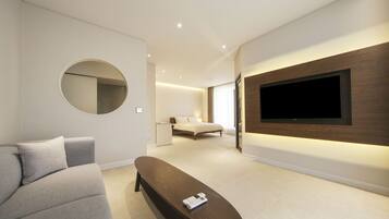 A modern hotel room with a large flat-screen TV mounted on the wall, a round mirror, a grey sofa, and a wooden bench.