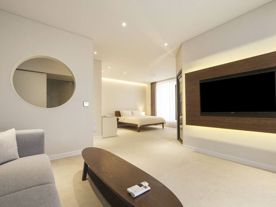 A modern hotel room with a large flat-screen TV mounted on the wall, a round mirror, a grey sofa, and a wooden bench.