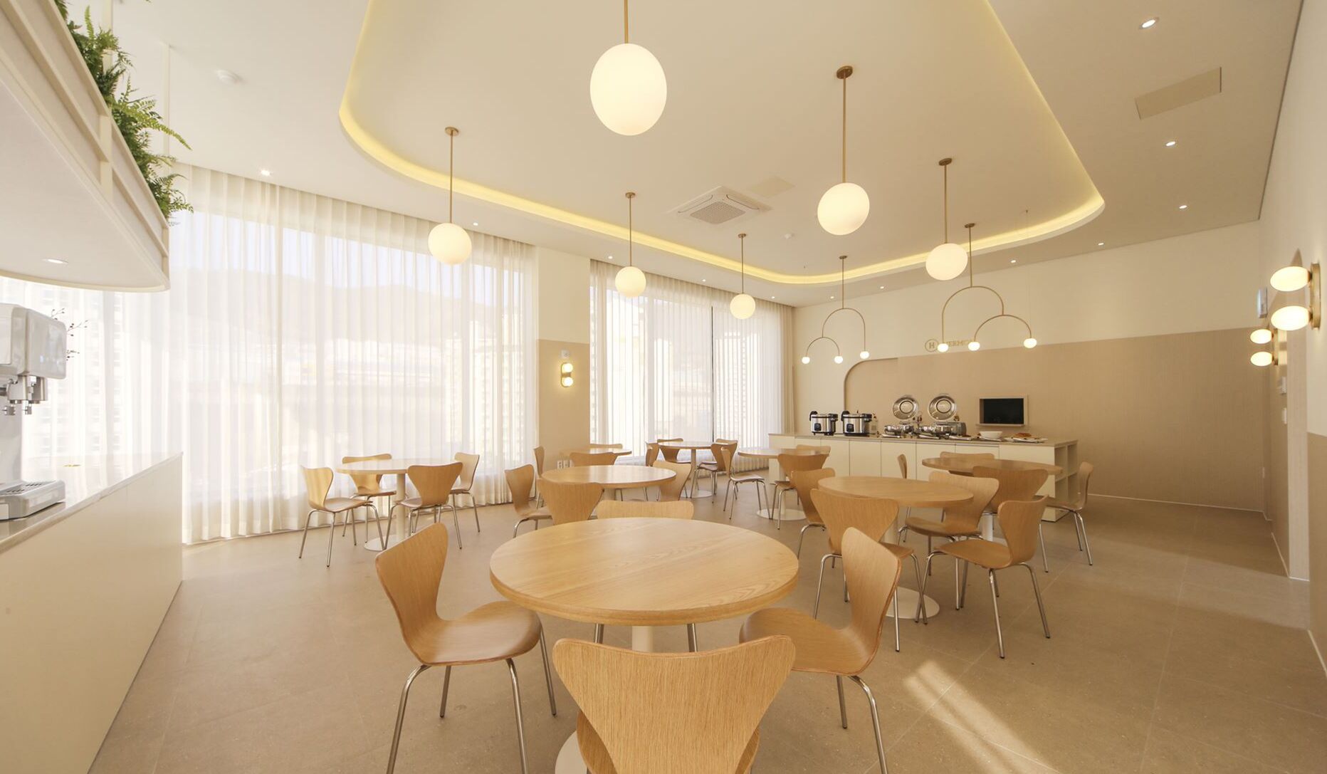 A modern dining area with round tables and chairs, large windows with sheer curtains, and pendant lighting.