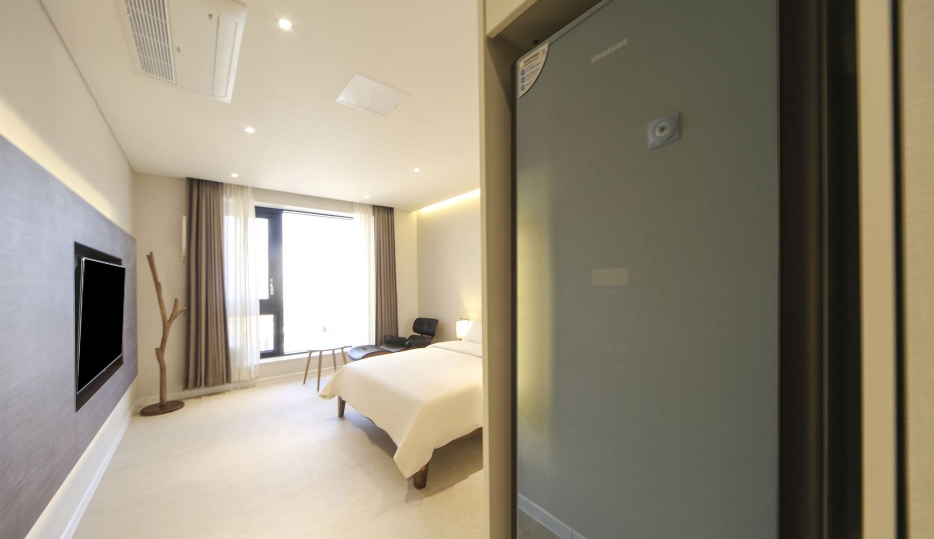A hotel room with a bed, a desk, a chair, a TV mounted on the wall, and a window with curtains.
