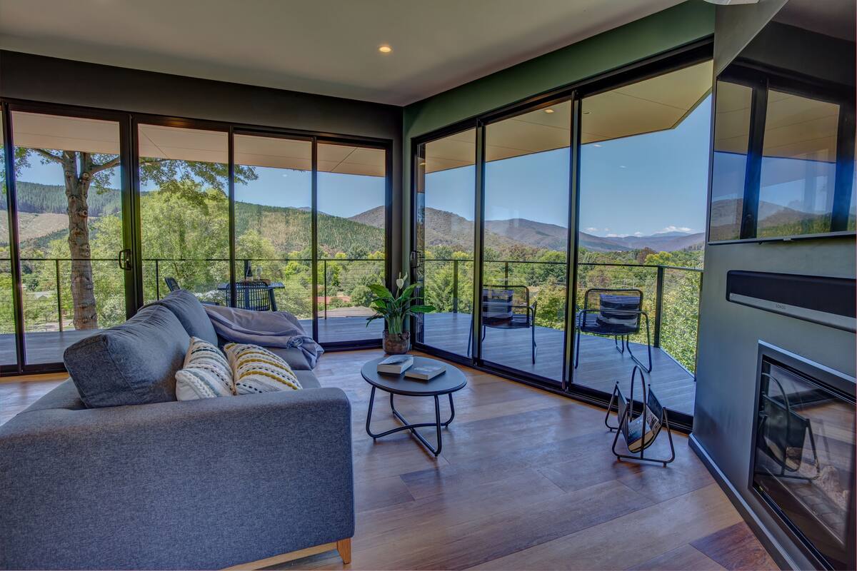Un salon moderne avec un canapé, une petite table et une cheminée. De grandes fenêtres offrent une vue sur l’extérieur.