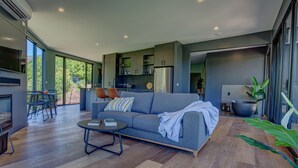 A modern living room with a grey sofa, a coffee table, and a TV. There is a kitchen area with cabinets and a refrigerator. Large windows provide natural light and a view of the outdoors. - The View Bright (Bright)