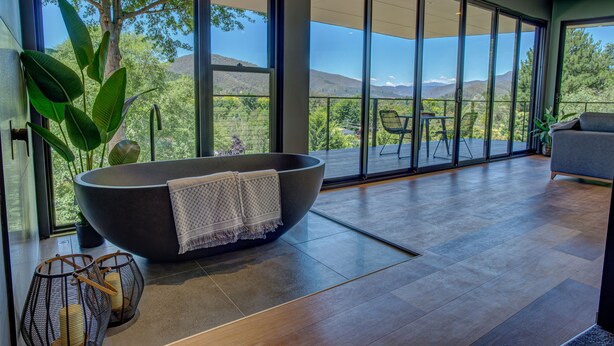 A modern bathroom with a large bathtub, a potted plant, and a view of the outdoors through large windows. - The View Bright (Bright)
