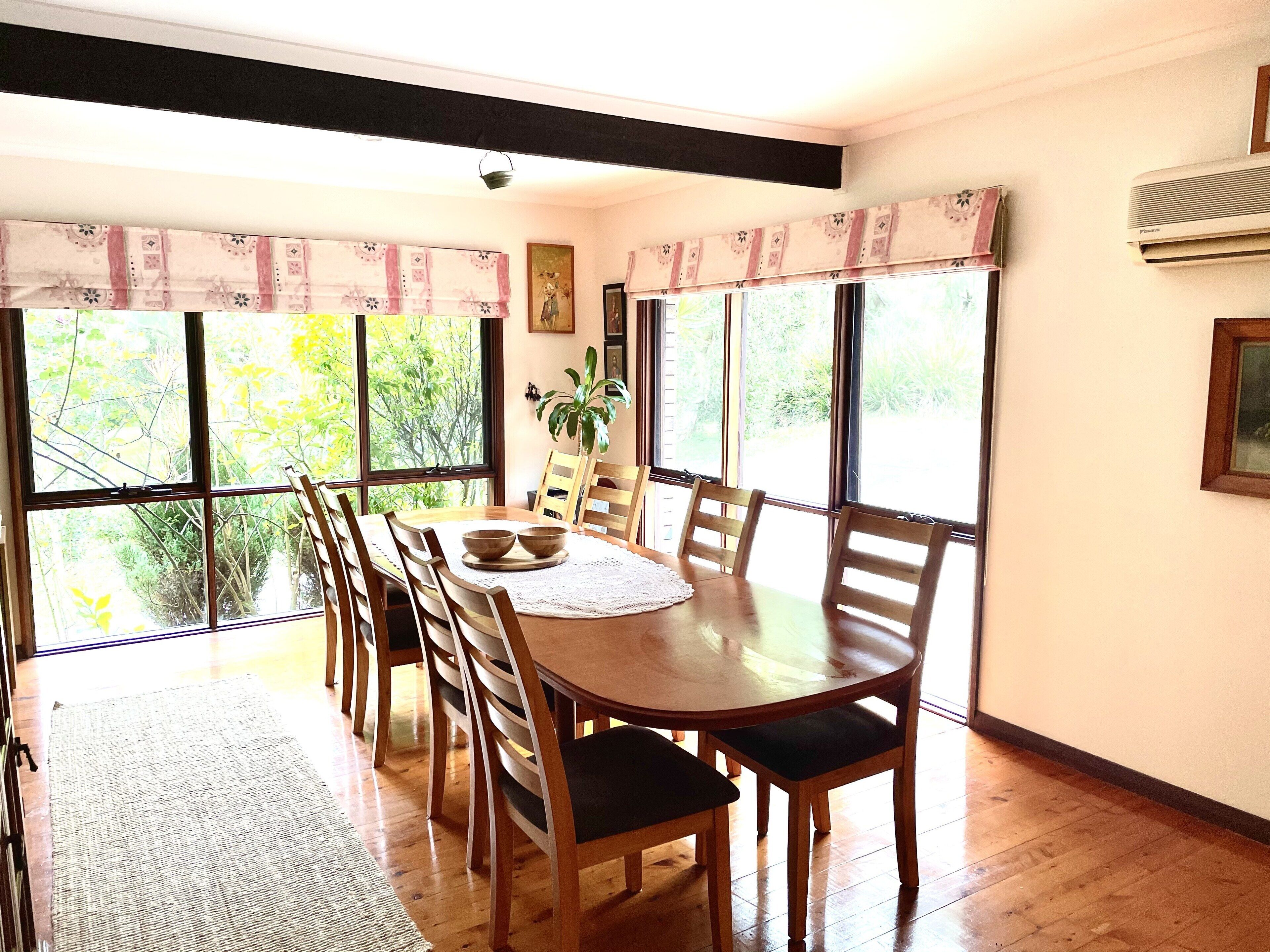 A dining area with a wooden table and chairs, large windows with curtains, a potted plant, and an air conditioning unit.