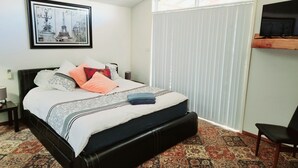 Ein Schlafzimmer mit einem Bett, einem Nachttisch, einem Sessel, einem Fenster mit Jalousien und einem am Wand montierten Fernseher.