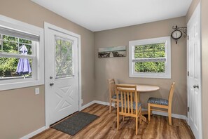 Une pièce avec une porte, deux fenêtres équipées de stores, une petite table avec des chaises, une horloge murale et un paillasson à l’entrée.