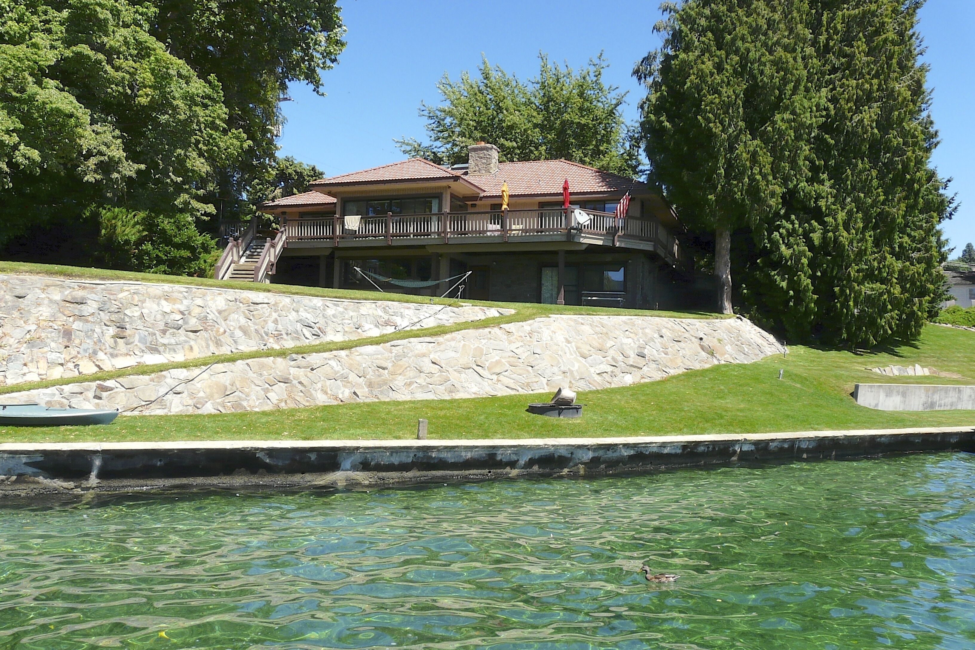 Une propriété au bord du lac, dotée d’un mur de soutènement en pierre, d’une terrasse en bois et offrant une vue sur l’eau.