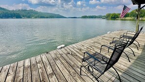 A wooden dock with metal chairs and an American flag, overlooking a calm lake with hills in the background.