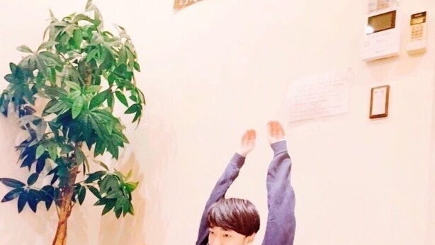 A person sitting on a couch with two poodles, one in front and one behind, in a room with a plant and decorative panels on the wall. - Polaris 101 (Nagoya)