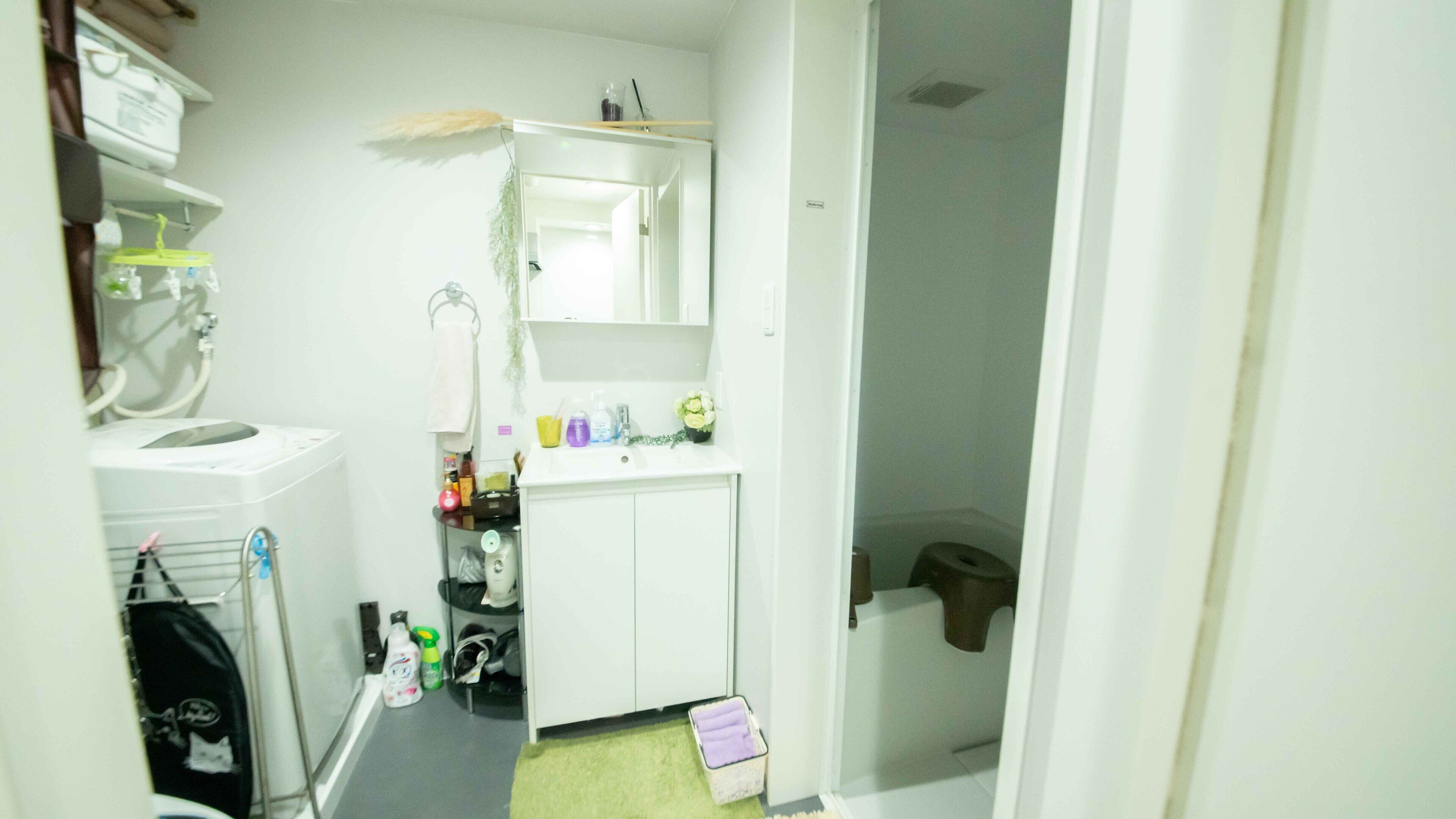 A small, compact bathroom with a white sink, mirror, and toilet. There is a shelf above the sink holding toiletries and a small plant. A towel hangs on the wall, and a trash bin is visible in the corner.