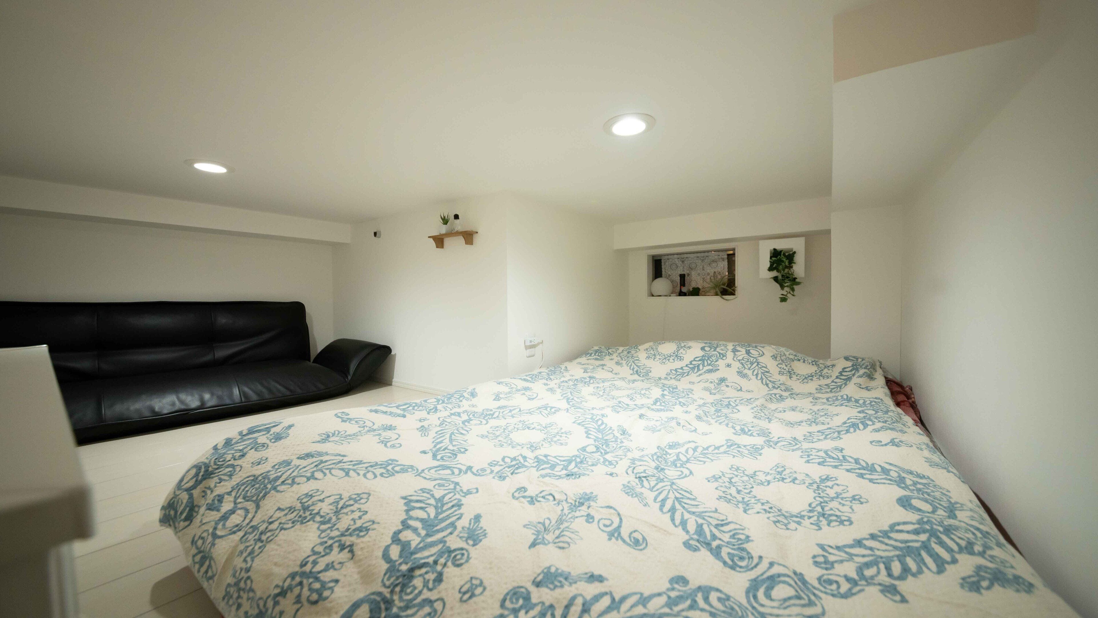 Ein Schlafzimmer mit einem Bett, einem schwarzen Ledersofa, einem kleinen Regal mit Pflanzen und einem Wandbild.