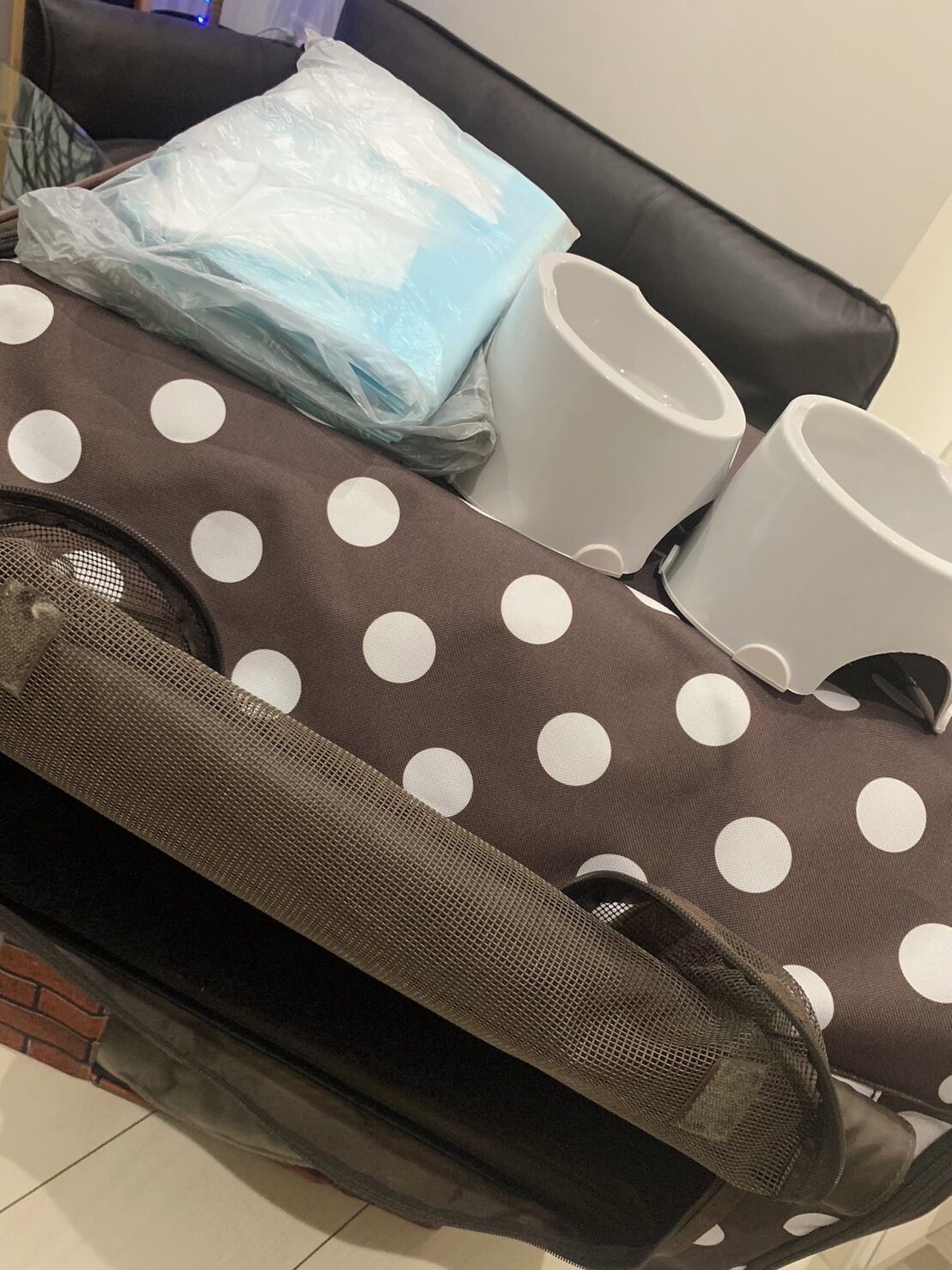A suitcase with a polka dot pattern, a plastic bag, and two white mugs on a black leather couch.
