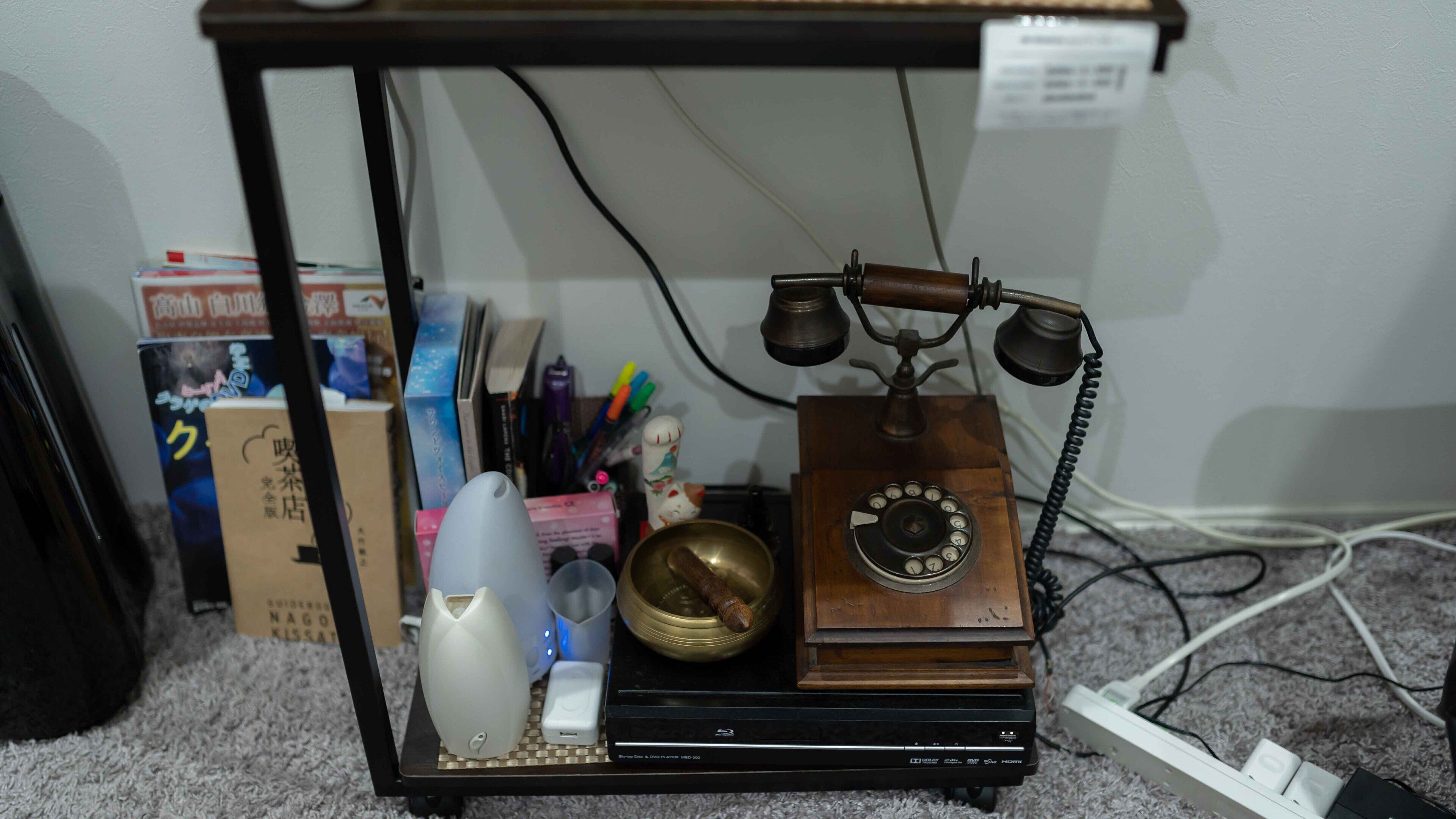En gammal telefon placerad på ett trästativ, tillsammans med en liten klocka, en metronom och några böcker.
